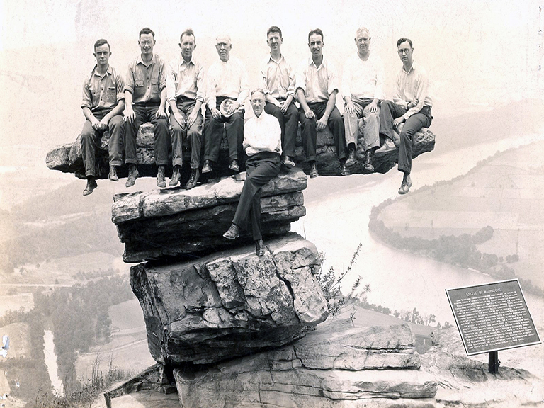 Nine Christian Brothers dressed in everyday clothing, are seated on a very precarious rock perched over the ledge of the Tennessee River valley down below.